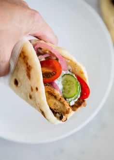 a hand is holding a pita sandwich on a white plate with cucumbers and tomatoes