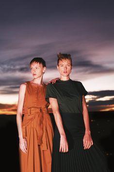 two women standing next to each other in front of a sky with clouds and the sun setting