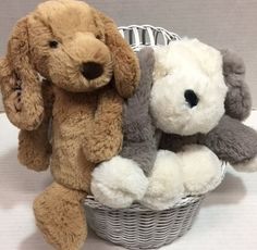 two stuffed dogs sitting in a basket on a white tableclothed surface, one is brown and the other is gray