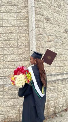 a woman in graduation gown holding flowers and a book on the side of a building