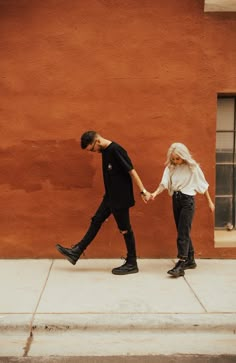 a man and woman holding hands while walking down the sidewalk in front of a building