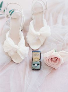 two pairs of shoes with bows on them and a ring in the middle, sitting next to a flower