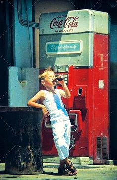 a young boy standing next to a coca cola machine talking on a cell phone and drinking from a cup