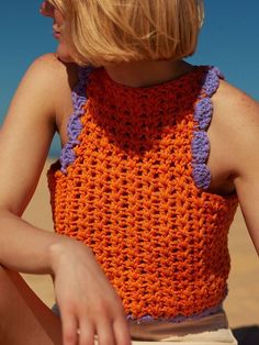 a woman wearing an orange and purple crochet top sitting in the sand with her hand on her hip