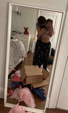 two people standing in front of a mirror with boxes on the floor and clothes all around them