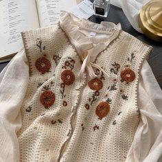 an open book on a table next to a white sweater with orange flowers and leaves