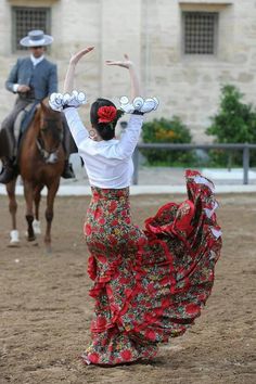 a woman in a long dress is dancing with her hands up while a man rides a horse behind her