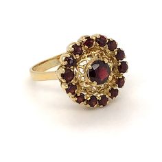 Yellow gold garnet cluster ring. The 18 karat yellow gold ring contains a halo of brownish red 2.5 mm round garnets surrounding a single round garnet that measures 5.5 mm in diameter. The halo surrounds a filigree pattern that separates the halo from the center stone. The top structure of halo and filigree is supported by a double gallery design set atop a plain shank that is split top and bottom and connected to the gallery for support. The ring is size 7. Gold Ruby Cluster Ring With Halo Design, Heirloom Style Round Garnet Cluster Ring, Heirloom Style Garnet Cluster Ring, Heirloom Garnet Cluster Ring, Heirloom Gold Cluster Ring With Garnet, Ruby Ring Designs, Split Top, Edwardian Jewelry, Filigree Pattern