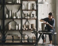 a woman sitting at a table in front of a shelf filled with vases