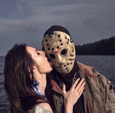 a woman kissing a man wearing a mask in front of the water with trees behind her