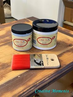 two jars of paint sitting on top of a wooden table next to a red brush