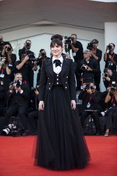 a woman in a long black dress standing on a red carpet with cameras around her