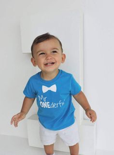 a little boy standing in front of a white wall wearing a blue shirt with the words my onederfit on it