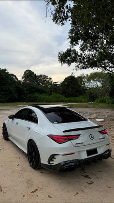 the rear end of a white car parked on top of a dirt field next to trees