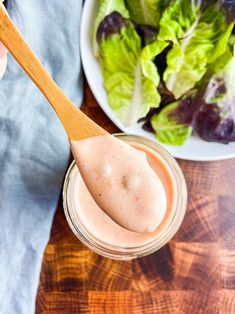 a person holding a wooden spoon over a jar of dressing next to a bowl of salad