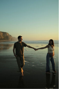 a man and woman holding hands while standing on the beach near the ocean at sunset