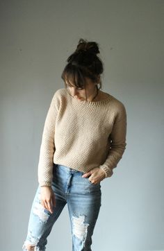 a woman in ripped jeans and a sweater stands against a white wall with her hands on her hips