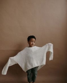 a little boy standing in front of a brown wall wearing a white sweater and black pants