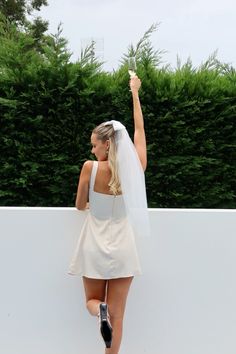 a woman in a short white dress is holding her hand up to the sky while wearing a veil