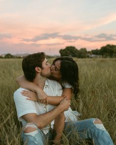 a man and woman sitting in tall grass kissing each other while the sun is setting