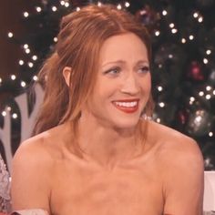 a woman sitting in front of a christmas tree smiling at the camera while talking to someone