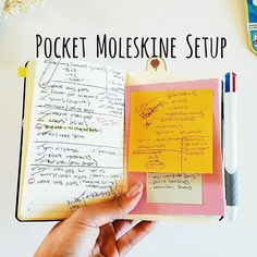 a hand holding an open notebook with notes and pens on it next to other items