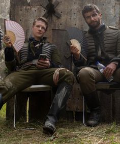 two men dressed in period clothing sit on benches holding fans and looking at their cell phones