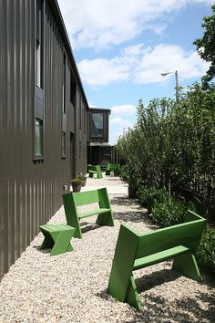 two green benches sitting next to each other in front of a building with trees and bushes