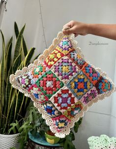 a crocheted pillow is being held by a woman's hand over a potted plant