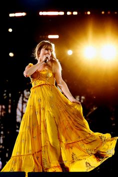 a woman in a yellow dress on stage with her hands up to her face and lights behind her