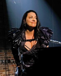 a woman in black dress standing next to a piano on stage with light coming from behind her