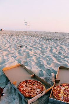 two pizzas are sitting in boxes on the sand at the beach, one is half eaten