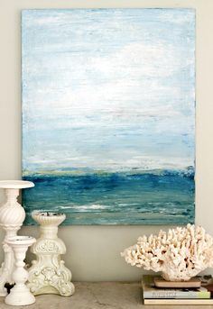 a white vase sitting on top of a table next to a painting with the ocean in it