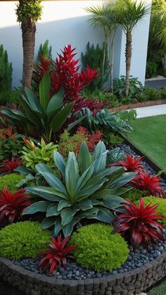 a garden filled with lots of green and red flowers next to a white wall on top of a lush green field