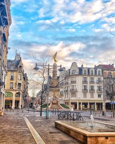 a city street with buildings and a fountain in the middle, surrounded by cobblestones