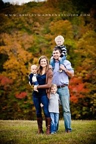 a family posing for a photo in front of fall trees with their toddler son on his shoulders