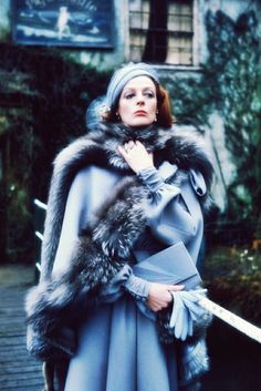 a woman in a blue dress and fur stoler standing on a wooden walkway with her hands under her chin