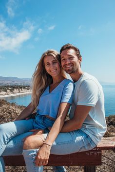 a man and woman sitting on top of a wooden bench next to the ocean in front of them