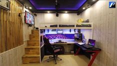 a computer desk sitting under a purple light in a room with wooden walls and stairs