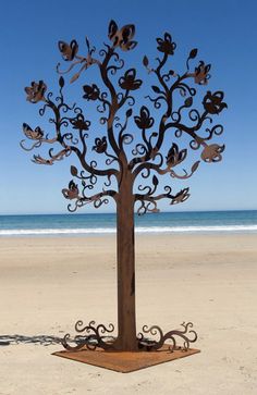 a metal tree sculpture sitting on top of a sandy beach next to the ocean and blue sky