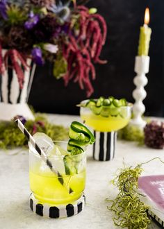 two glasses filled with cucumber and lemonade sitting on a table next to flowers