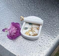 a white box with some jewelry on it next to a purple flower and water droplets