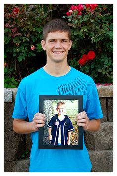 a young man holding up a framed photo in front of some bushes and flowers with red roses behind him