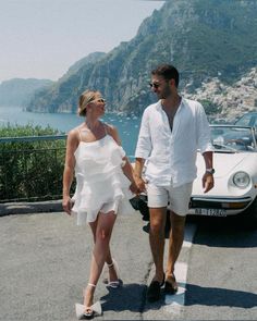 a man and woman holding hands while walking next to a car on the side of a road