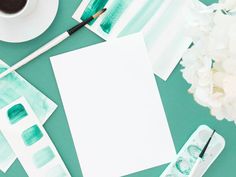 watercolor paints and paper on a green table with white flowers, paintbrushes and cup of coffee