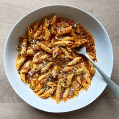 a white bowl filled with pasta and sauce on top of a wooden table next to a spoon