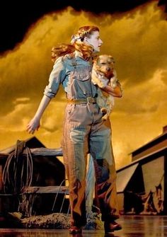 a woman in overalls holding a dog and standing on a wooden platform with stormy skies behind her