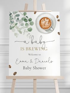 a baby is brewing welcome sign sitting on top of an easel next to a cup of coffee