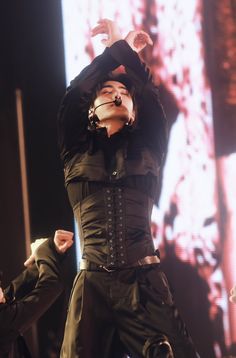 a man in black performing on stage with his hands up to the air and one arm raised