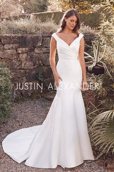 a woman in a white wedding dress standing next to a stone wall and plants with her hands on her hips